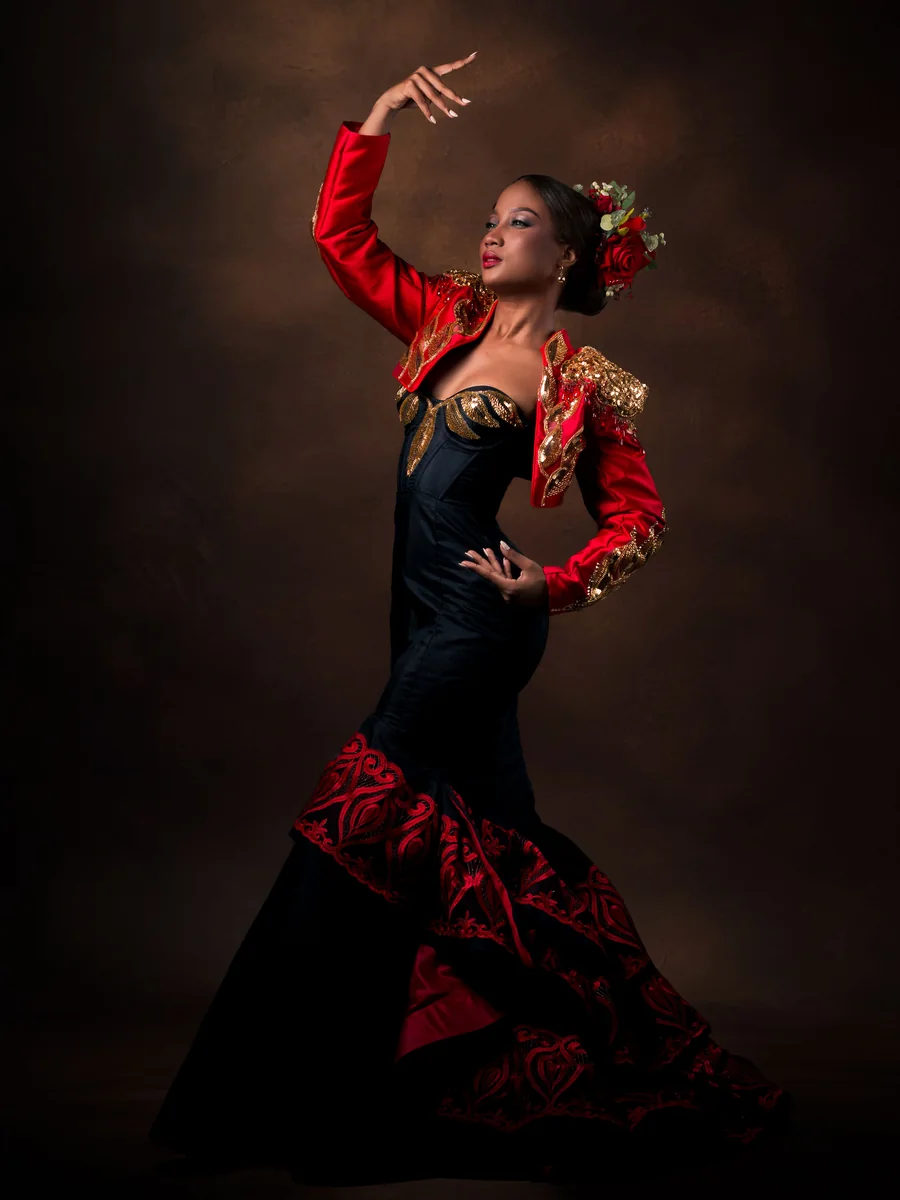 Tancerka flamenco w czerwono-czarnej sukni w połowie pozowania, z jedną ręką uniesioną, na ciemnym tle studyjnym (zdjęcie poglądowe, nie przedstawia własnego zespołu Corral de la Morería)